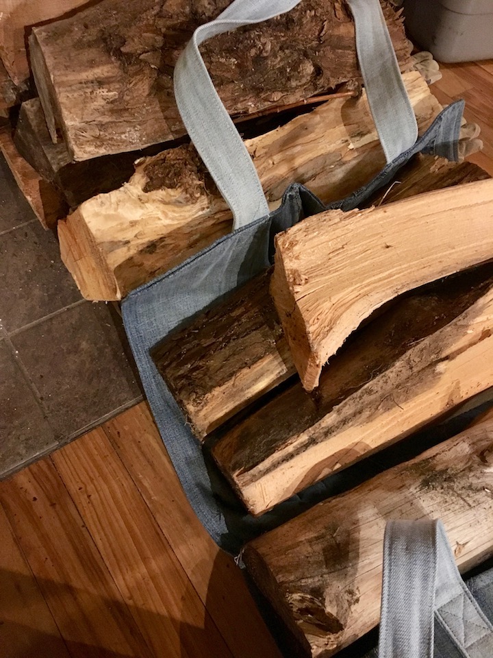 Pieces of firewood are stacked on top of a denim firewood sling, which is next to more wood stacked on the floor near a hearth.